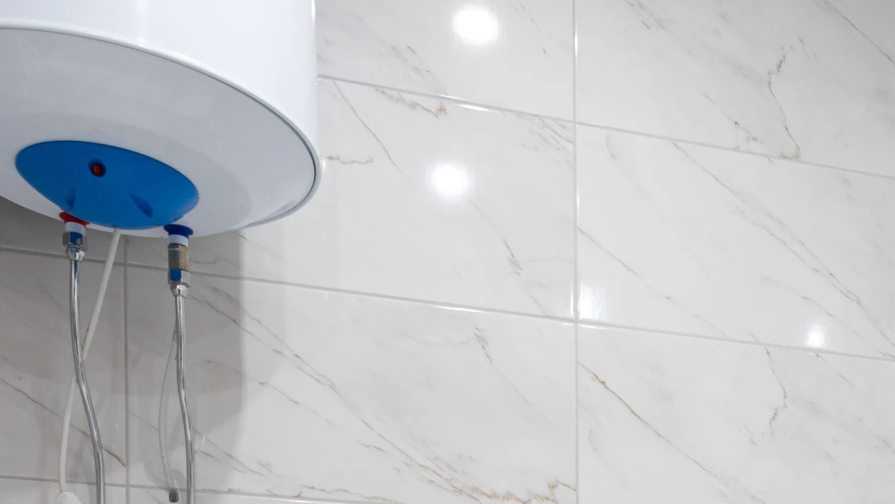 Underside of a wall-mounted electric water heater showing its blue cap and two flexible connection hoses against marble bathroom tiling