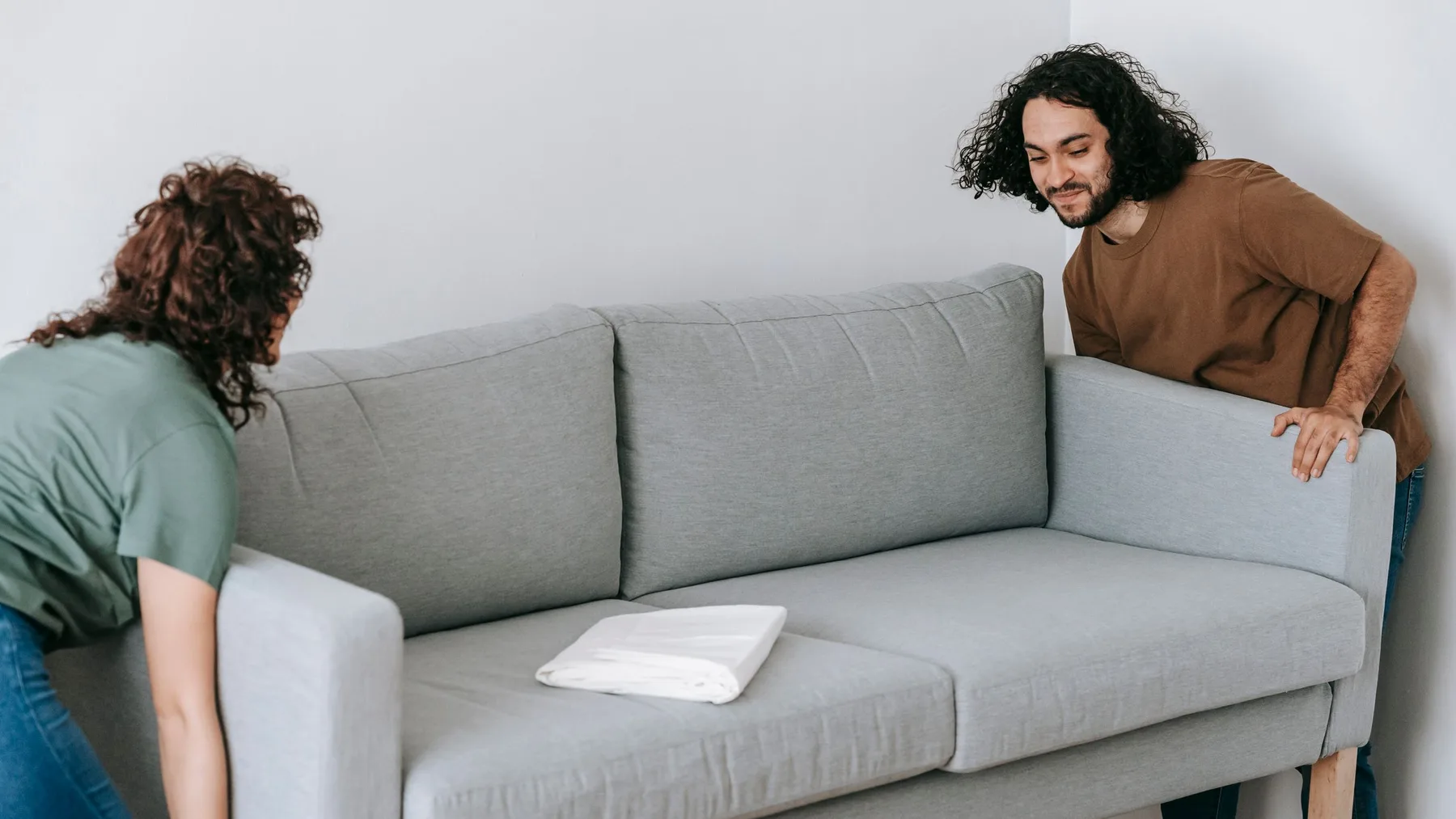 Two people lifting a grey three-seater fabric sofa away from a plain white wall in an empty room