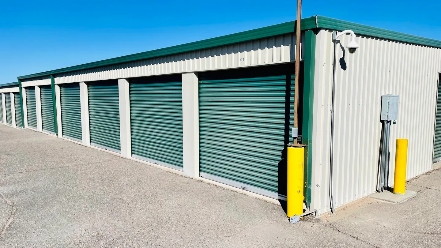 Row of drive-up self-storage units with green roller shutters and yellow safety bollards under a clear sky